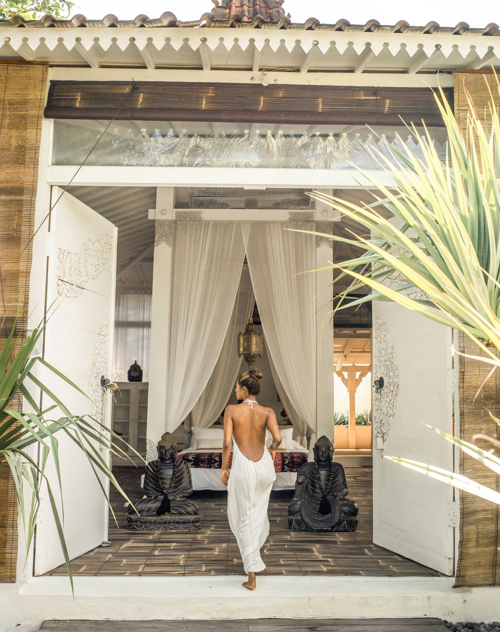 Woman in a white dress walking towards a tropical villa in  Bali with plants and statues.