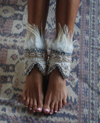 Feet wearing ornate cream ankle cuffs with feathers on a patterned fabric background