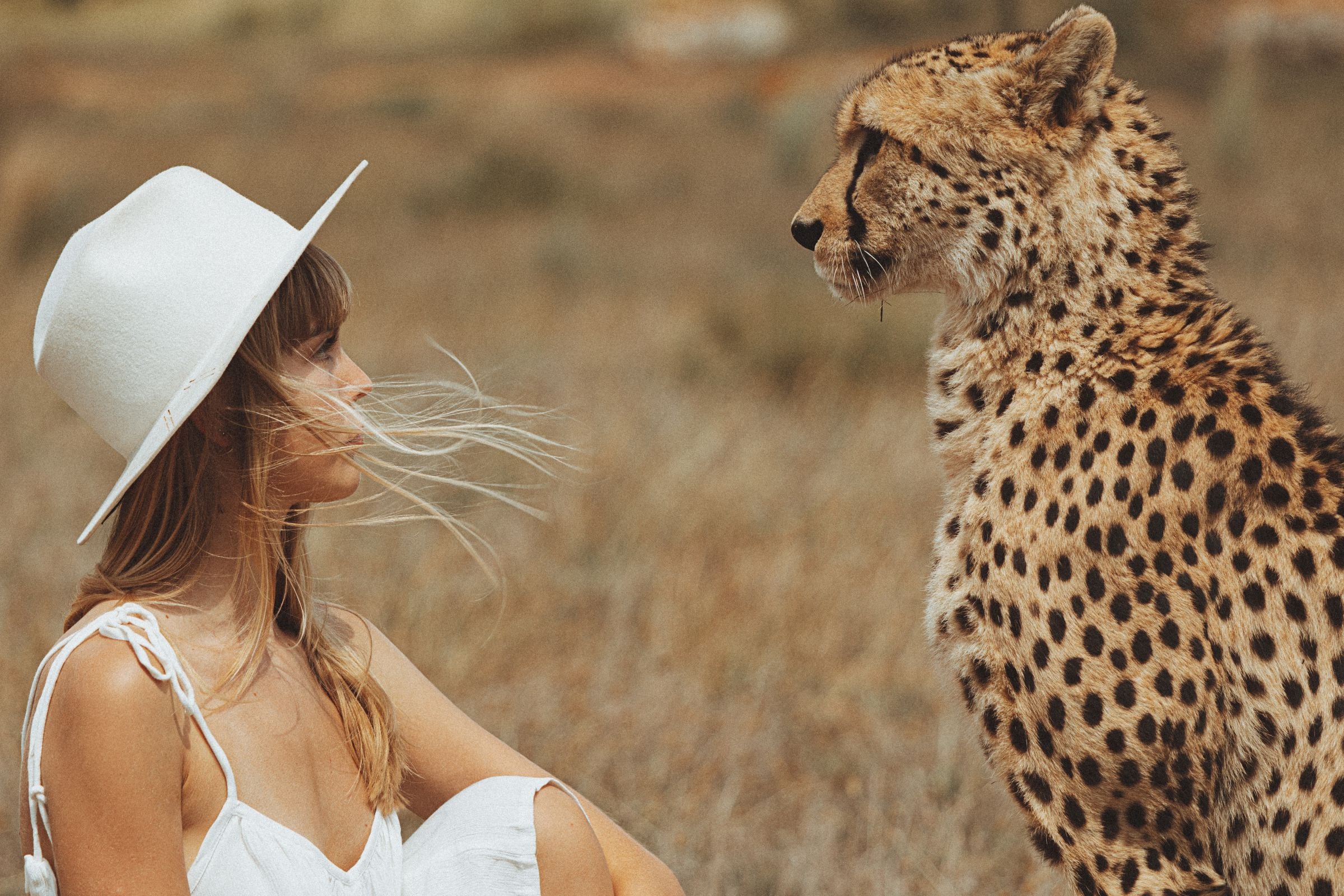 Woman in a white hat and dress sitting next to a cheetah in a natural setting