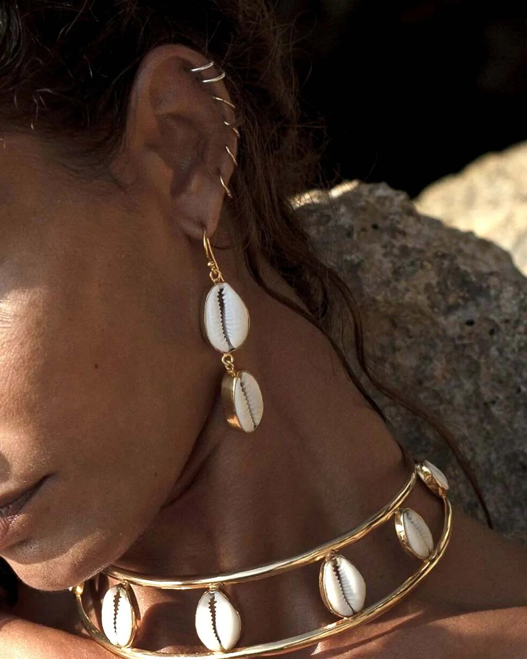 Close-up of a person wearing gold earrings and a gold bracelet with shell designs against a rocky background.