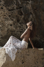 Person wearing a white crochet lace garment sitting against a rocky wall.