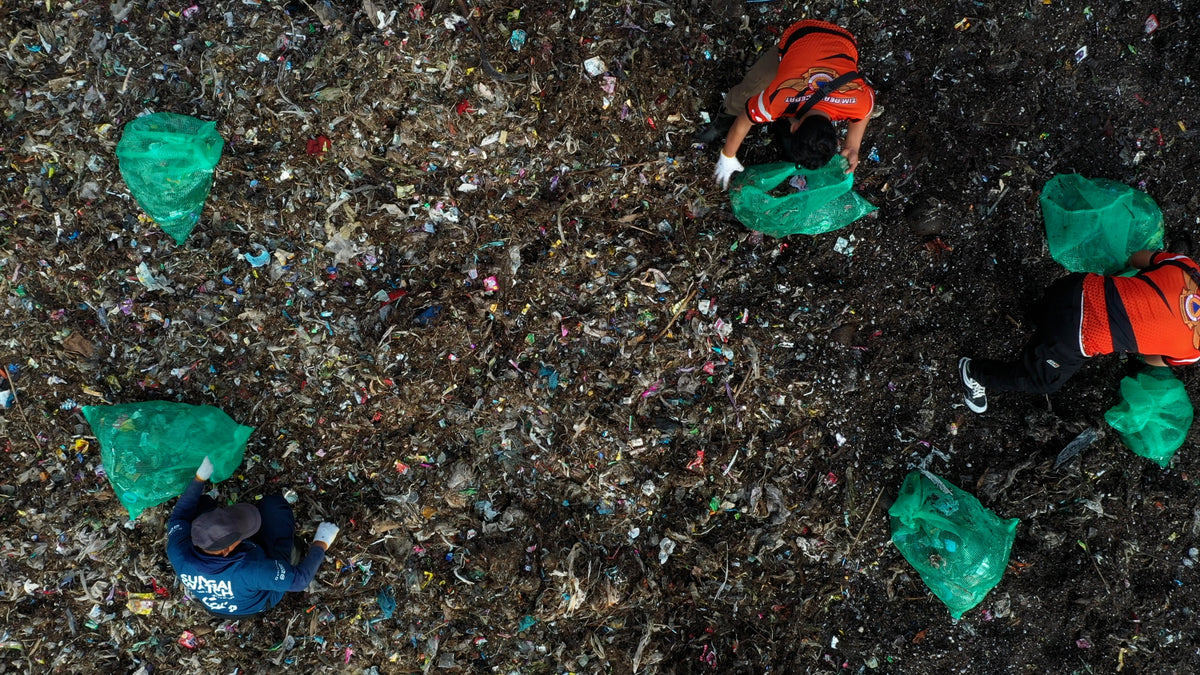 Small figures in orange vests and green bags amidst trash on a dark surface