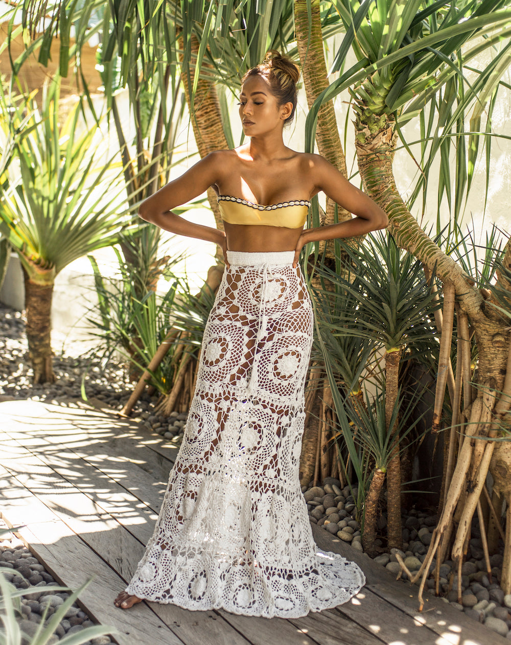 Woman in a gold bikini top and white crochet lace skirt standing in a tropical setting with palm trees.