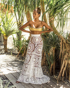 Woman in a gold bikini top and white crochet lace skirt standing in a tropical setting with palm trees.