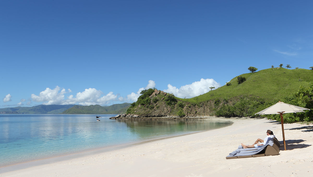 Person lounging on a beach with clear blue water and green hills in the background