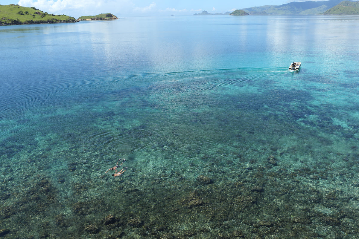 Clear blue water with a small boat and people in the distance, surrounded by green hills.