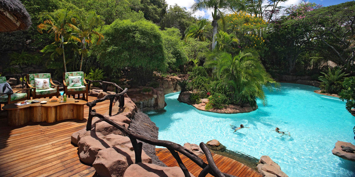 Pool area with lounge chairs, tables, and a natural rock formation surrounded by trees.