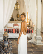 Woman in a white backless dress standing in a bedroom with a Buddha statue and draped curtains.